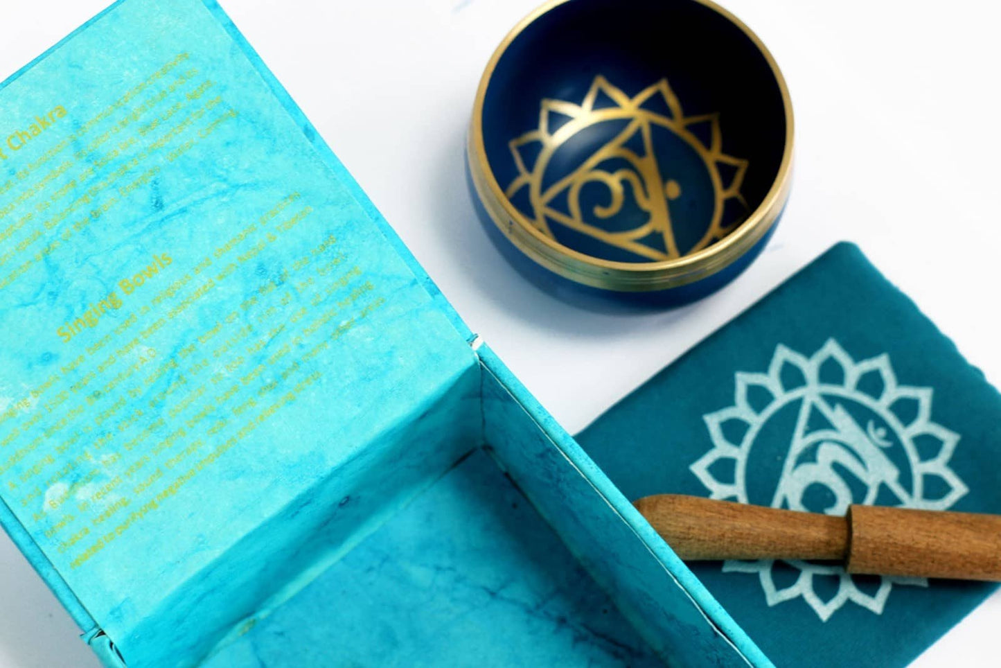 Coffret ouvert bleu turquoise contenant un bol tibétain bleu turquoise avec une bordure dorée. À l'intérieur, le symbole du Chakra Gorge doré. À côté, un tapis bleu turquoise avec le symbole du Chakra Gorge doré, et un fuseau en bois.