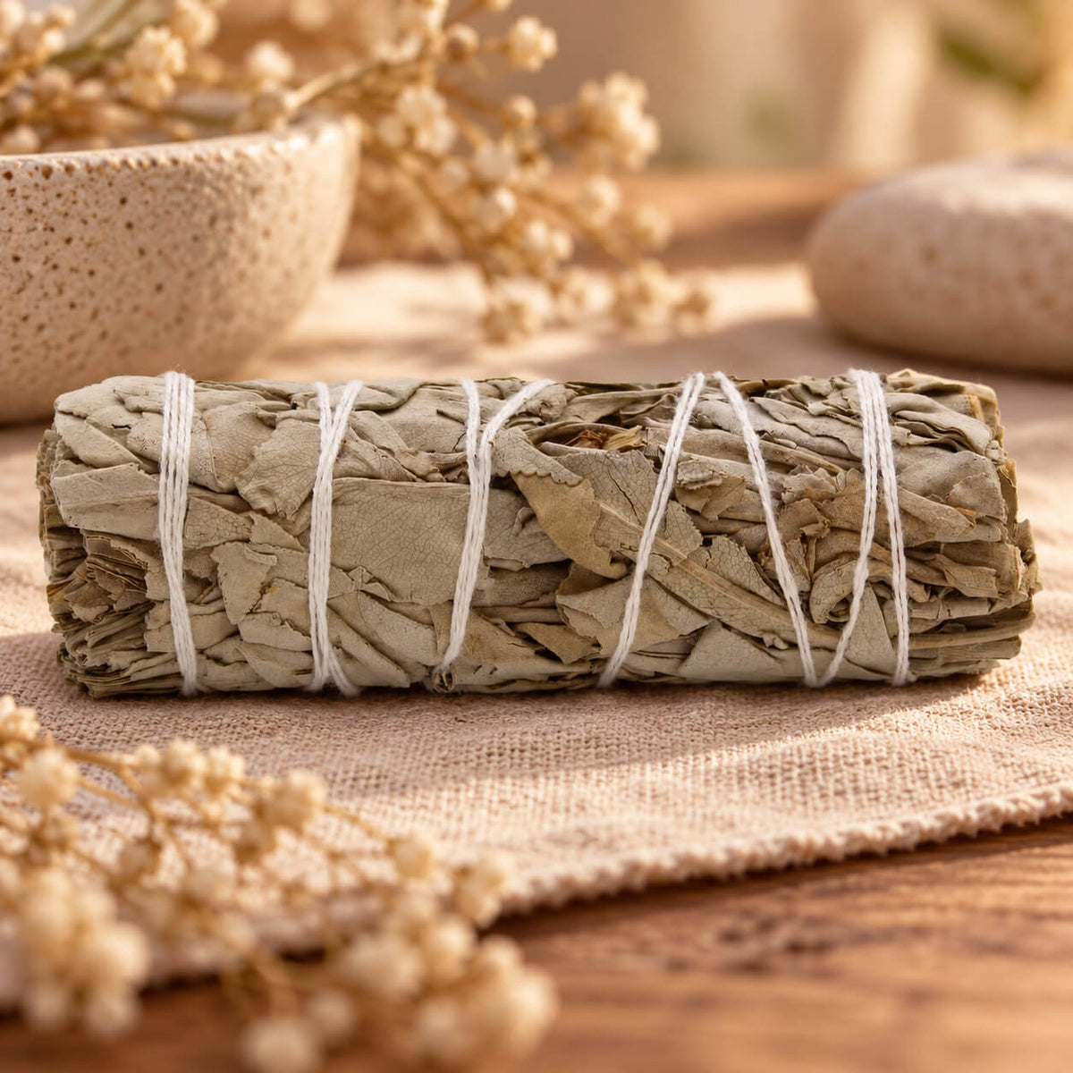 Bâton de sauge blanche nature lié de fil blanc, posé sur une étoffe beige près de fleurs séchées