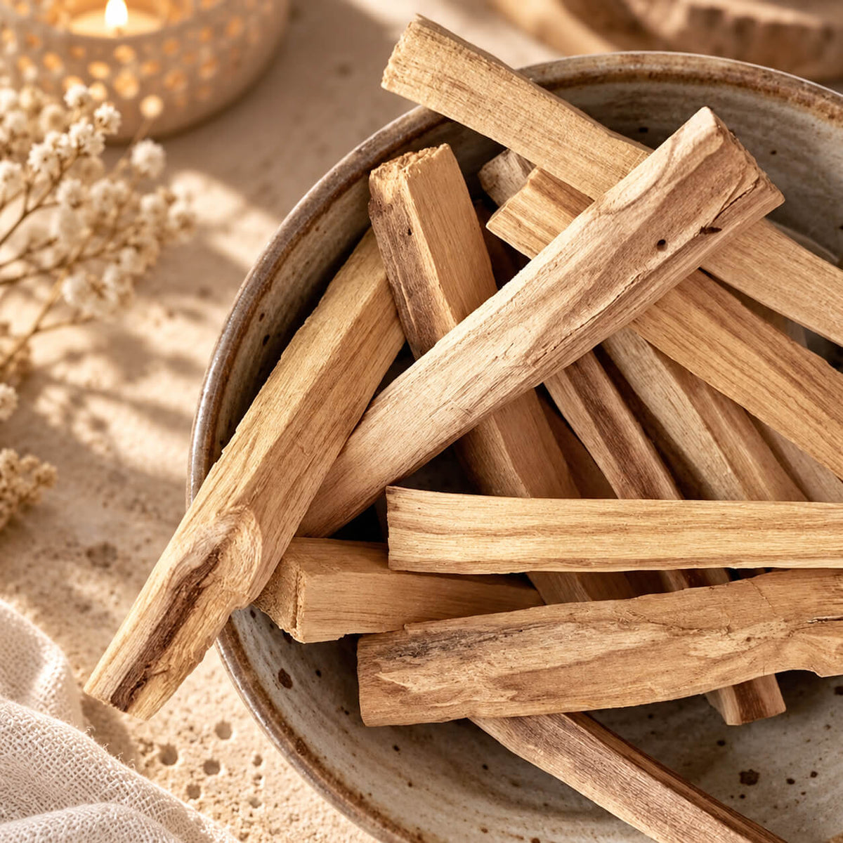 Bâtons de palo santo en bois blond veiné, empilés dans un bol en céramique près d'une bougie