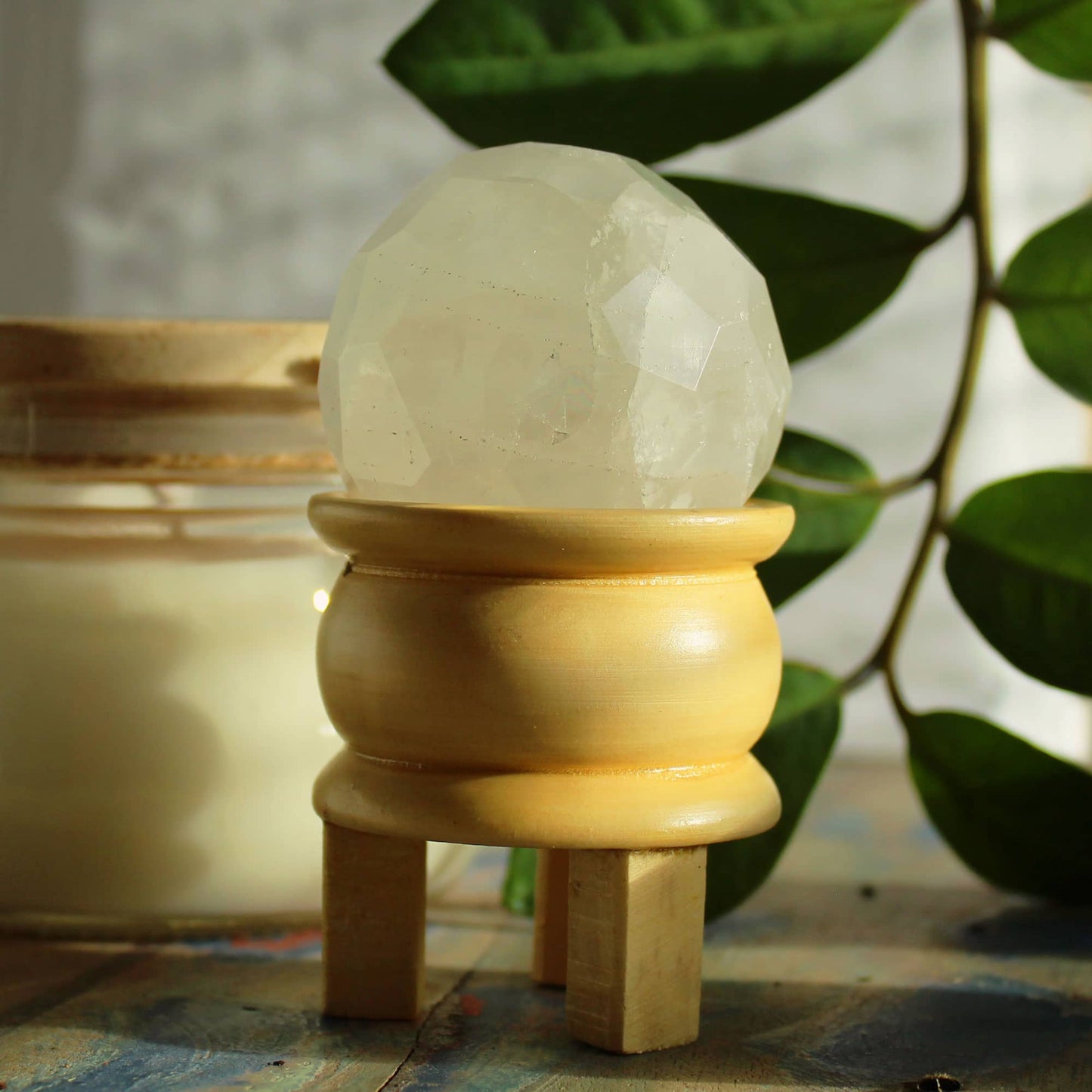 Sphère facettée en Quartz Blanc cristallin aux reflets purs et lumineux, montée sur un support en bois clair, mise en scène avec feuillage vert.