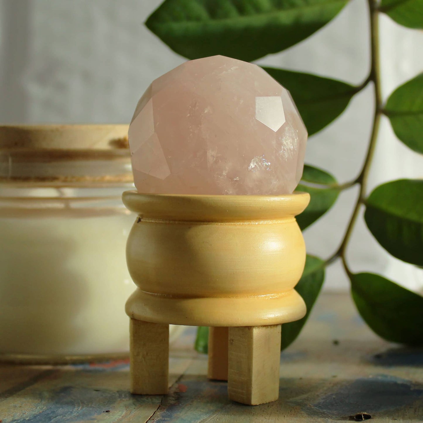 Sphère facettée en Quartz Rose aux reflets doux et rosés, montée sur un support en bois clair, mise en scène avec feuillage vert.