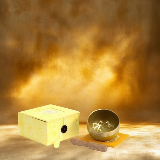 Coffret fermé jaune contenant un bol tibétain jaune avec une bordure dorée. À l'intérieur, le symbole du Chakra Plexus Solaire doré, posé sur un tapis jaune, accompagné d'un fuseau en bois sur fond jaune solaire.