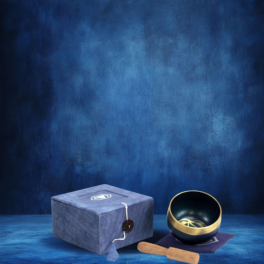 Coffret fermé bleu foncé à côté, un bol tibétain bleu foncé avec une bordure dorée. À l'intérieur, le symbole du Chakra Troisième Œil doré, posé sur un tapis bleu foncé, accompagné d'un fuseau en bois sur fond bleu.