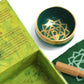 Coffret ouvert vert contenant un bol tibétain vert avec une bordure dorée. À l'intérieur, le symbole du Chakra Cœur doré. À côté, un tapis vert avec le symbole du Chakra Cœur doré, et un fuseau en bois.