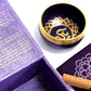 Coffret ouvert violet contenant un bol tibétain violet avec une bordure dorée et le symbole du Chakra Couronne doré à l'intérieur. À côté, un tapis violet avec le symbole du Chakra Couronne doré, et un fuseau en bois.