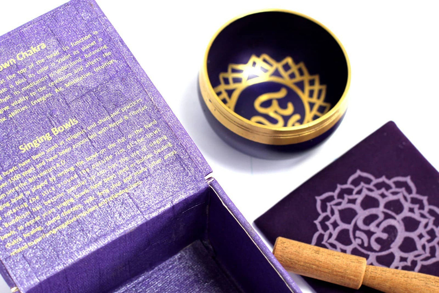 Coffret ouvert violet contenant un bol tibétain violet avec une bordure dorée et le symbole du Chakra Couronne doré à l'intérieur. À côté, un tapis violet avec le symbole du Chakra Couronne doré, et un fuseau en bois.