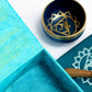 Coffret ouvert bleu turquoise contenant un bol tibétain bleu turquoise avec une bordure dorée. À l'intérieur, le symbole du Chakra Gorge doré. À côté, un tapis bleu turquoise avec le symbole du Chakra Gorge doré, et un fuseau en bois.