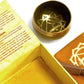 Coffret ouvert jaune contenant un bol tibétain jaune avec une bordure dorée. À l'intérieur, le symbole du Chakra Plexus Solaire doré. À côté, un tapis jaune avec le symbole du Chakra Plexus Solaire doré, et un fuseau en bois.