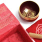 Coffret ouvert rouge contenant un bol tibétain rouge avec une bordure dorée. À l'intérieur, le symbole du Chakra Racine doré. À côté, un tapis rouge avec le symbole du Chakra Racine doré, et un fuseau en bois.