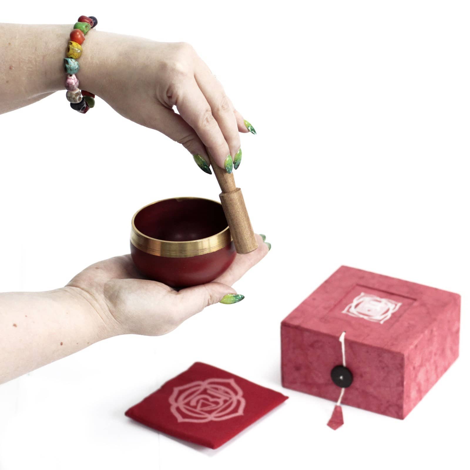 Coffret fermé rouge avec un tapis rouge et le symbole du Chakra Racine doré. À côté, une main tient un fuseau et un bol tibétain rouge avec une bordure dorée, prêt à être utilisé pour faire chanter le bol.