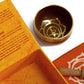 Coffret ouvert orange contenant un bol tibétain orange avec une bordure dorée. À l'intérieur, le symbole du Chakra Sacré doré. À côté, un tapis orange avec le symbole du Chakra Sacré doré, et un fuseau en bois.