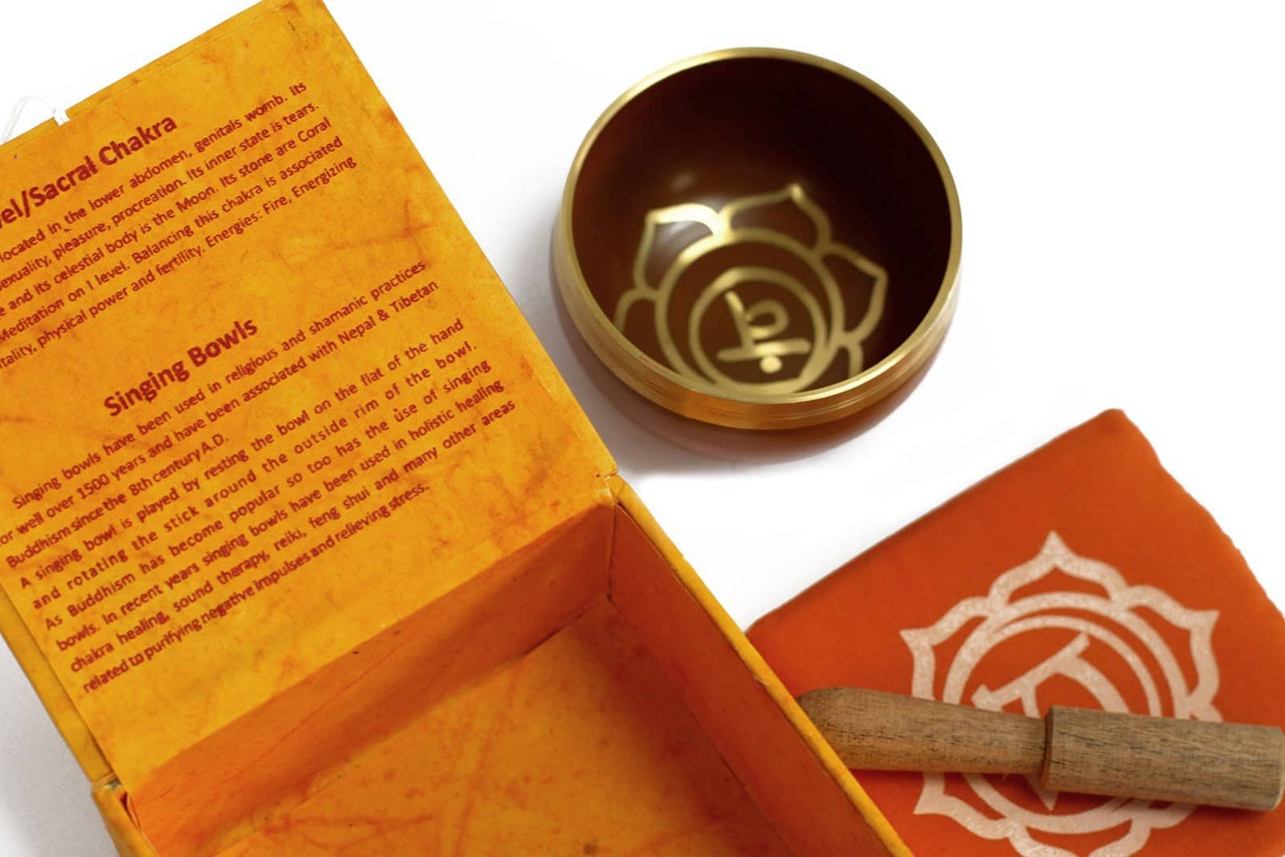 Coffret ouvert orange contenant un bol tibétain orange avec une bordure dorée. À l'intérieur, le symbole du Chakra Sacré doré. À côté, un tapis orange avec le symbole du Chakra Sacré doré, et un fuseau en bois.