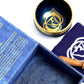 Coffret ouvert bleu foncé contenant un bol tibétain bleu foncé avec une bordure dorée. À l'intérieur, le symbole du Chakra Troisième Œil doré. À côté, un tapis bleu foncé avec le symbole du Chakra Troisième Œil doré, et un fuseau en bois.