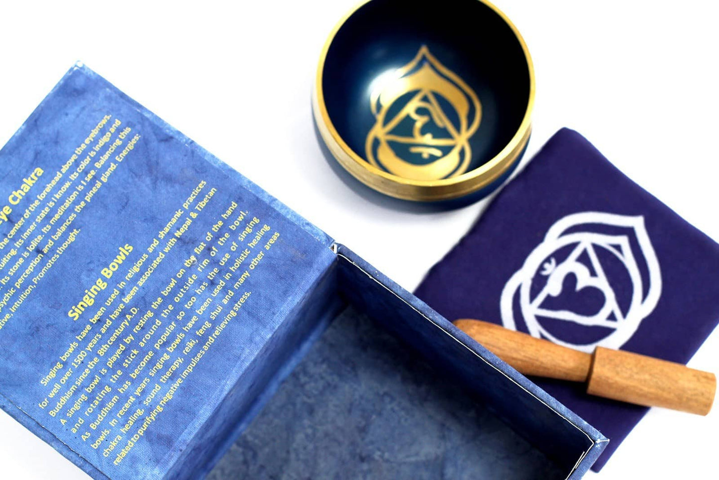 Coffret ouvert bleu foncé contenant un bol tibétain bleu foncé avec une bordure dorée. À l'intérieur, le symbole du Chakra Troisième Œil doré. À côté, un tapis bleu foncé avec le symbole du Chakra Troisième Œil doré, et un fuseau en bois.