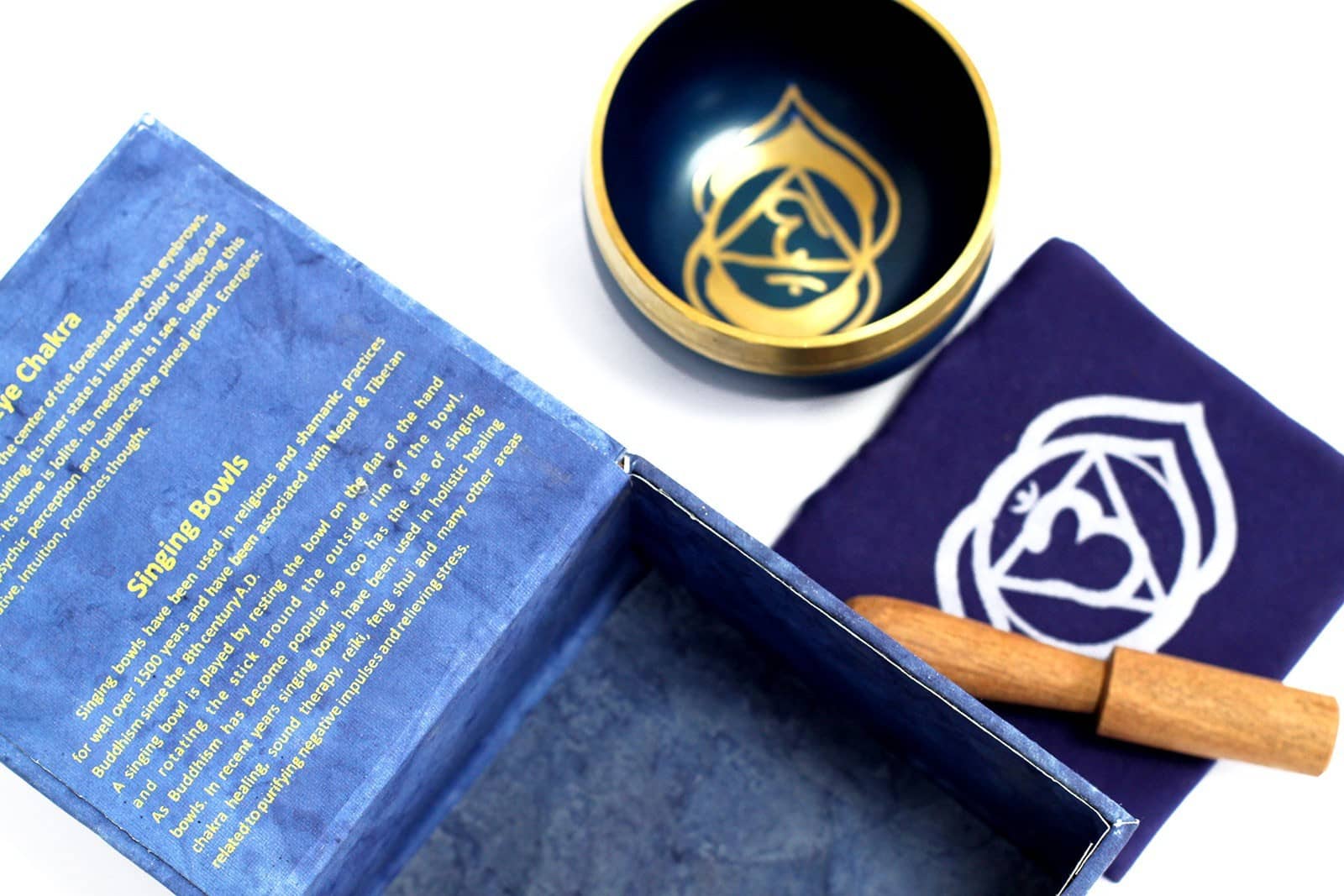 Coffret ouvert bleu foncé contenant un bol tibétain bleu foncé avec une bordure dorée. À l'intérieur, le symbole du Chakra Troisième Œil doré. À côté, un tapis bleu foncé avec le symbole du Chakra Troisième Œil doré, et un fuseau en bois.
