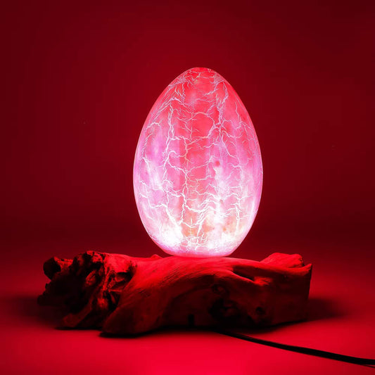 Lampe œuf rose craquelé allumée sur souche de bois flotté avec ambiance rouge dramatique.