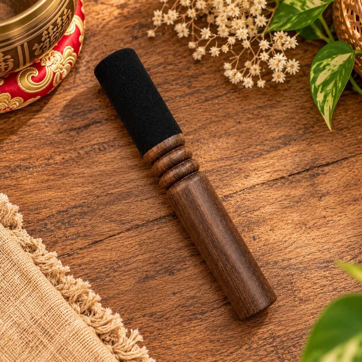 Bâton pour bol tibétain à manche droit en bois, trois anneaux tournés et embout gainé de noir, posé sur une table en bois près d’un bol sur coussin rouge