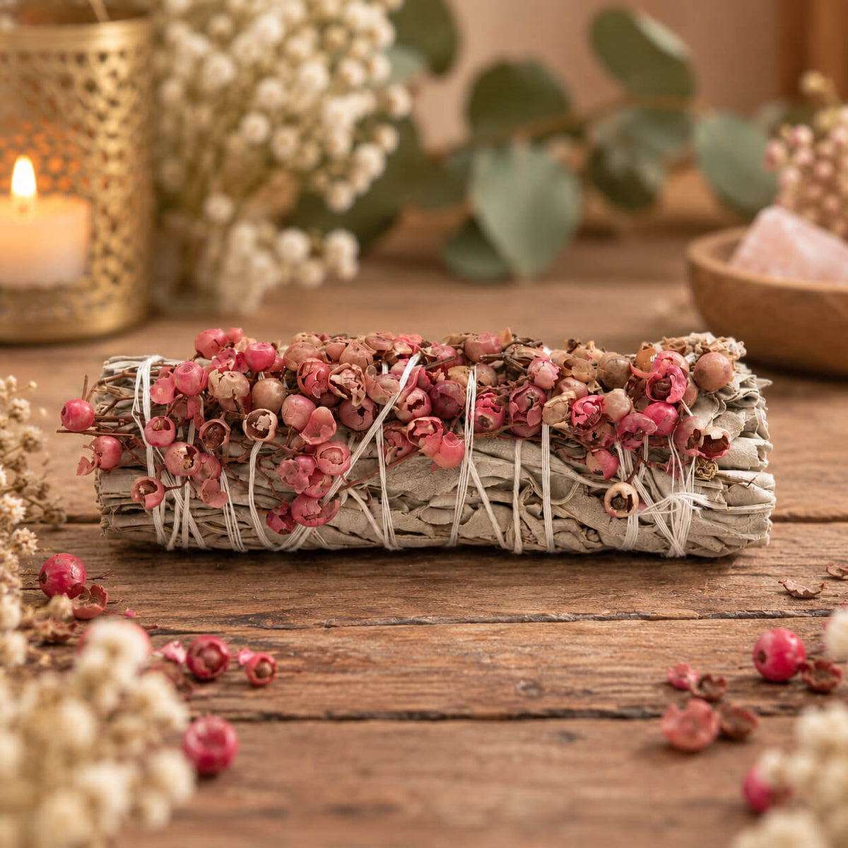 Bâton de sauge blanche couvert de grappes de baies de pirul roses et crème, posé sur une table en bois près d'une bougie