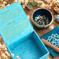 Symbole doré du chakra de la gorge au fond du bol bleu, près de la boîte turquoise ouverte et du coussin imprimé