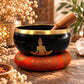 Bol chantant noir orné d'un Bouddha doré, posé sur un coussin rouge orangé avec son bâton en bois