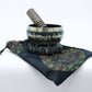 Un bol tibétain noir avec des bordures dorées et des mantras dorés gravés, posé sur un coussin qui est lui-même placé sur un sac assorti.