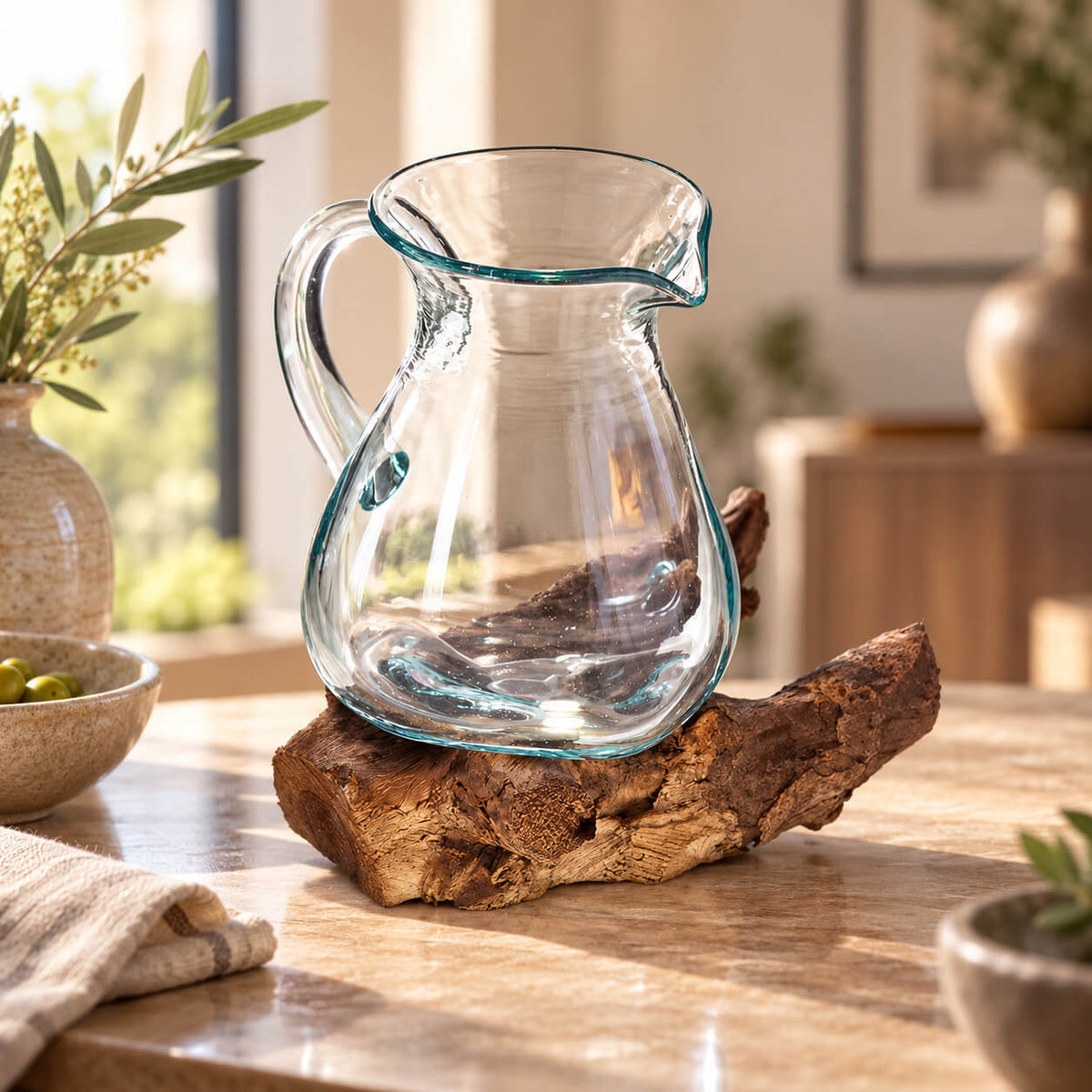 Carafe en verre soufflé sur socle en bois brut, avec une branche d'olivier en arrière plan