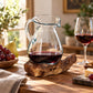 Carafe en verre soufflé sur socle en bois remplie de vin rouge, avec un plateau de fromage et de raisins