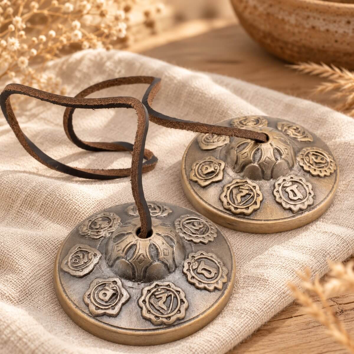 Paire de cymbales tingsha en laiton patiné, dôme en lotus et sept chakras en relief, posées sur du lin avec leur lanière en cuir