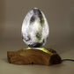 Lampe œuf de lave gris anthracite allumée révélant ses inclusions lumineuses sur socle bois flotté.