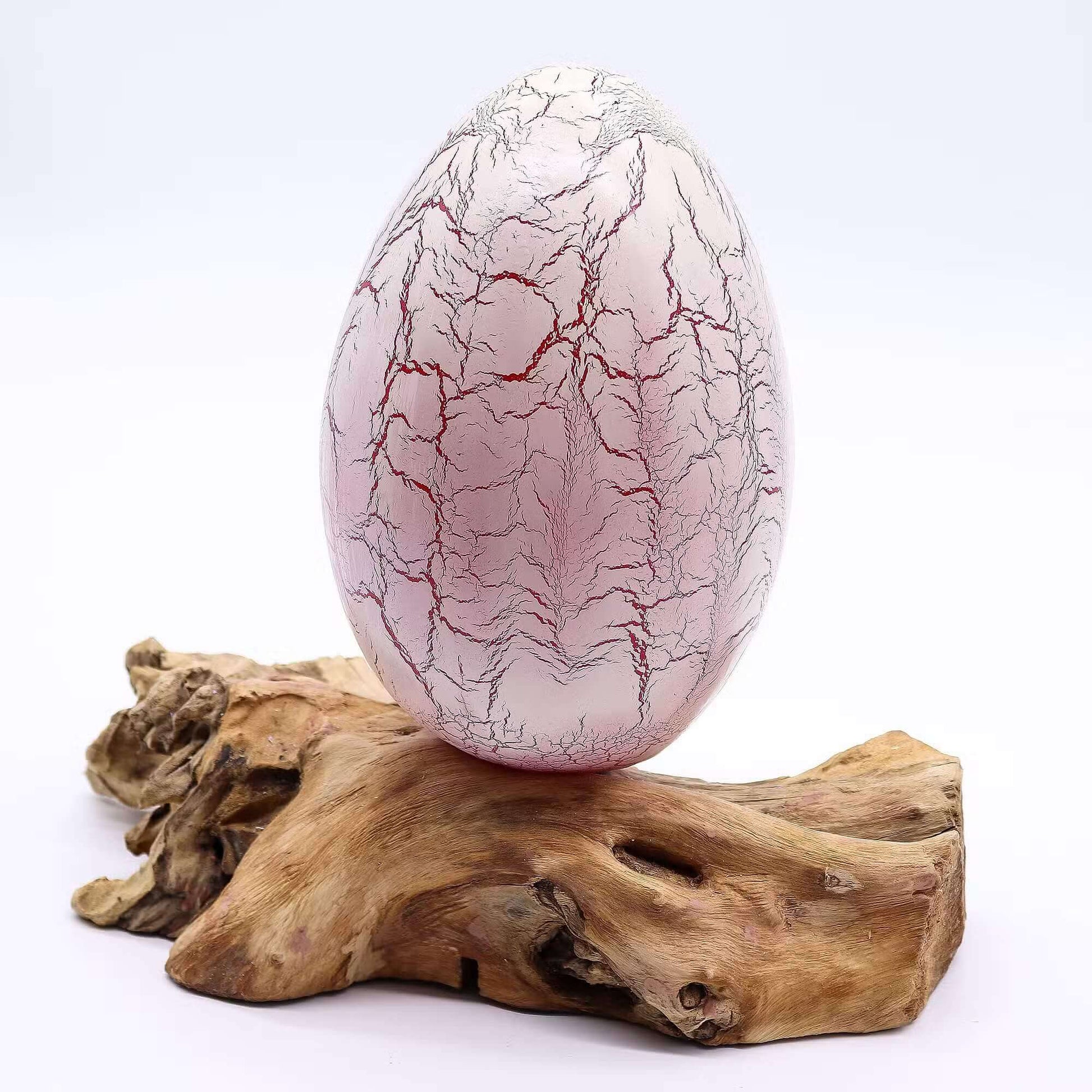 Lampe œuf rose poudré craquelé éteinte posée sur socle bois flotté brut sur fond blanc.