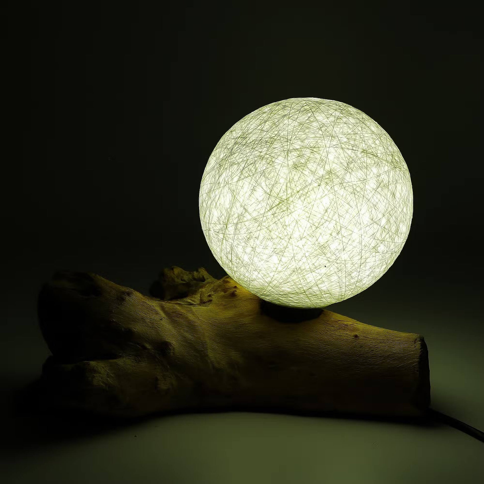 Lampe sphère lune ivoire allumée diffusant un halo doré sur branche de bois flotté.