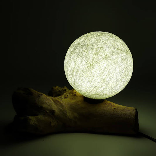 Lampe sphère lune ivoire allumée diffusant un halo doré sur branche de bois flotté.