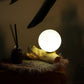 Lampe sphère lune ivoire allumée en décoration zen avec diffuseur sur meuble en bois.