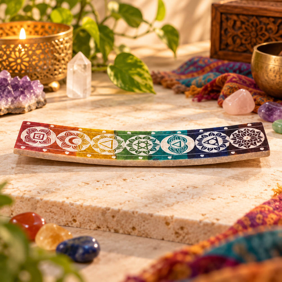Plateau porte-encens incurvé en stéatite gravé des sept chakras, en dégradé du rouge au violet, posé entre des pierres roulées