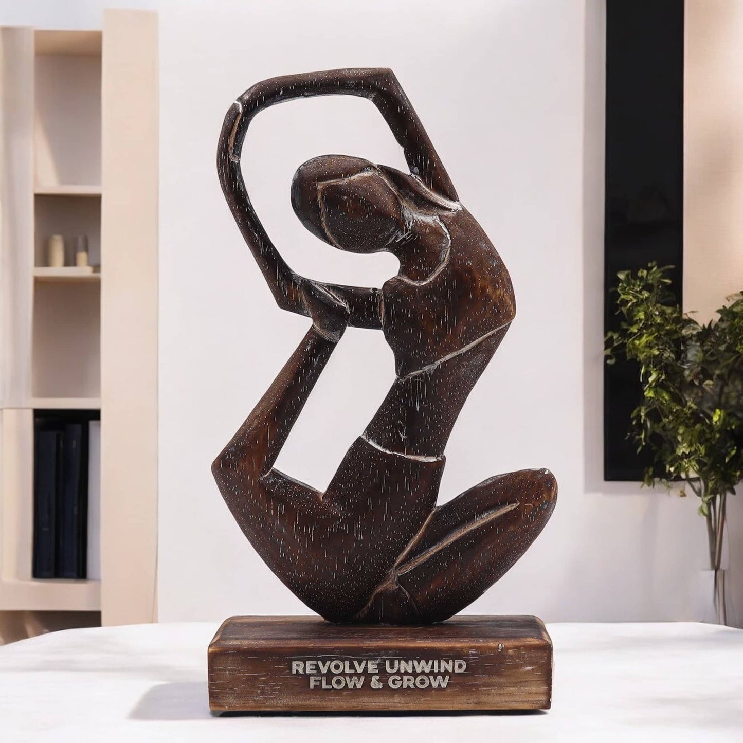 Statue de yoga en bois foncé représentant une pose de torsion fluide, sur socle gravé du mantra 'REVOLVE UNWIND FLOW & GROW', photographiée dans un intérieur épuré