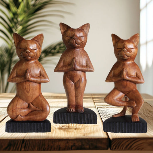 Trois modèles de chats en bois pratiquant différentes positions de yoga : lotus, salutation et arbre, chacun présenté sur un socle noir avec plante verte floue en arrière-plan