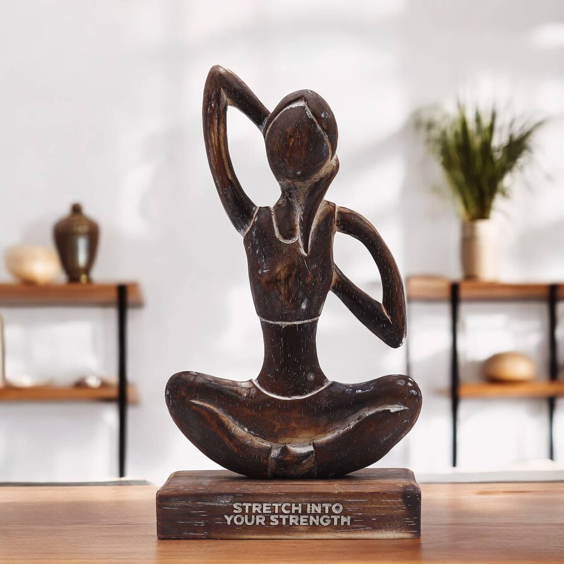 Statue de yoga en bois foncé représentant une posture d'étirement latéral, sur socle gravé du mantra 'STRETCH INTO YOUR STRENGTH', photographiée dans un décor minimaliste avec étagères en bois.