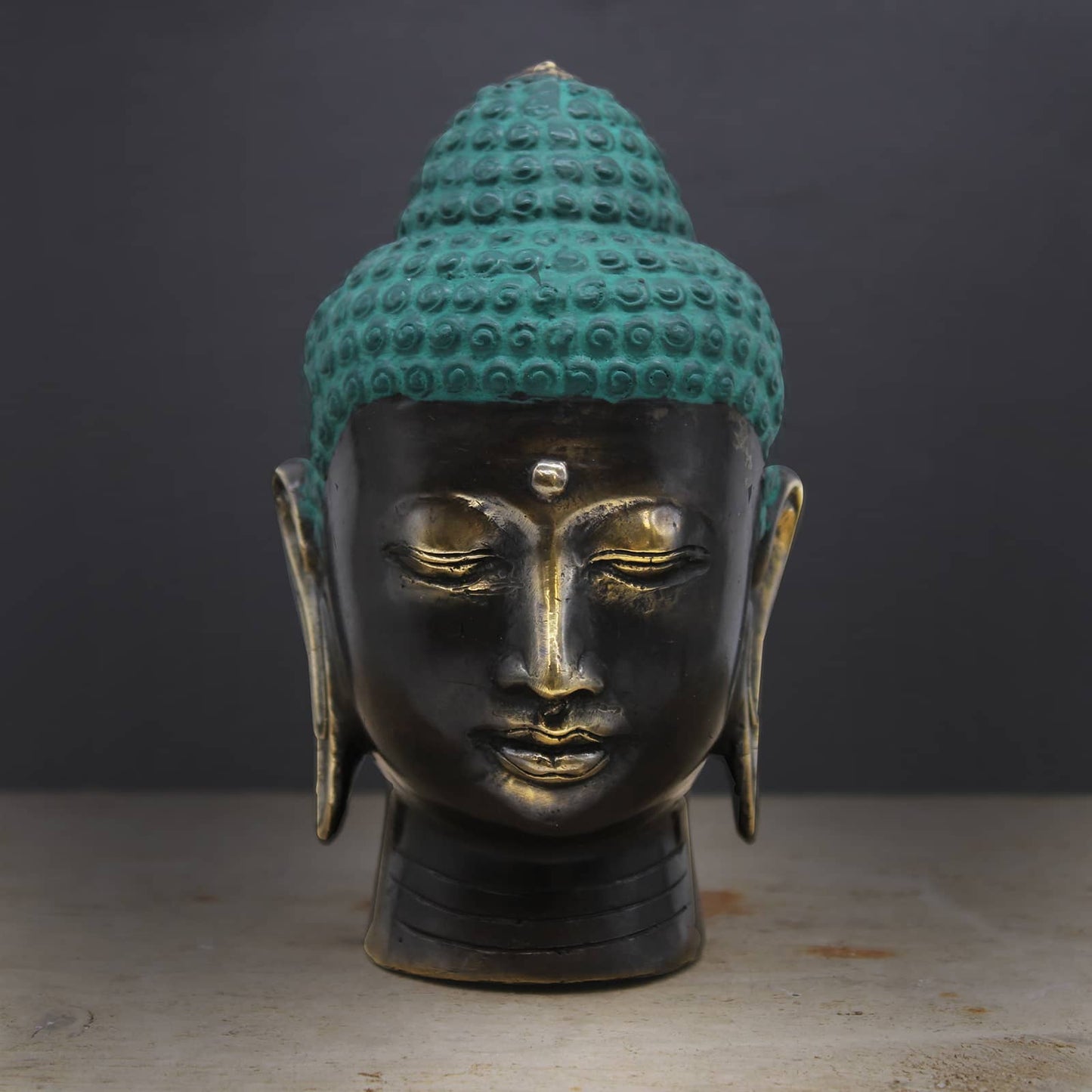 Tête de Bouddha en laiton à la coiffe turquoise patinée et au visage bronze antique, expression sereine sur fond sombre.