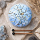 Tongue drum 18cm bleu ciel veiné d'or avec symbole Om et deux mailloches en bois sur pierre naturelle.