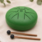 Tongue drum 16cm vert émeraude avec symbole Om gravé et deux mailloches en bois posées sur tissu écru.