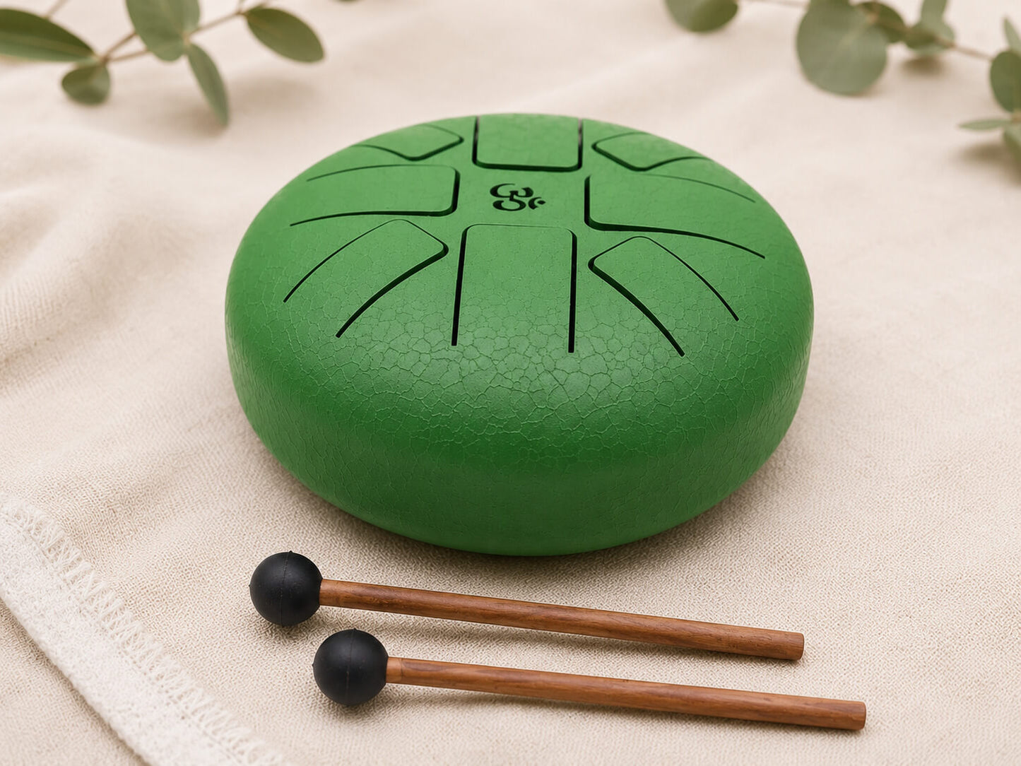 Tongue drum 16cm vert émeraude avec symbole Om gravé et deux mailloches en bois posées sur tissu écru.