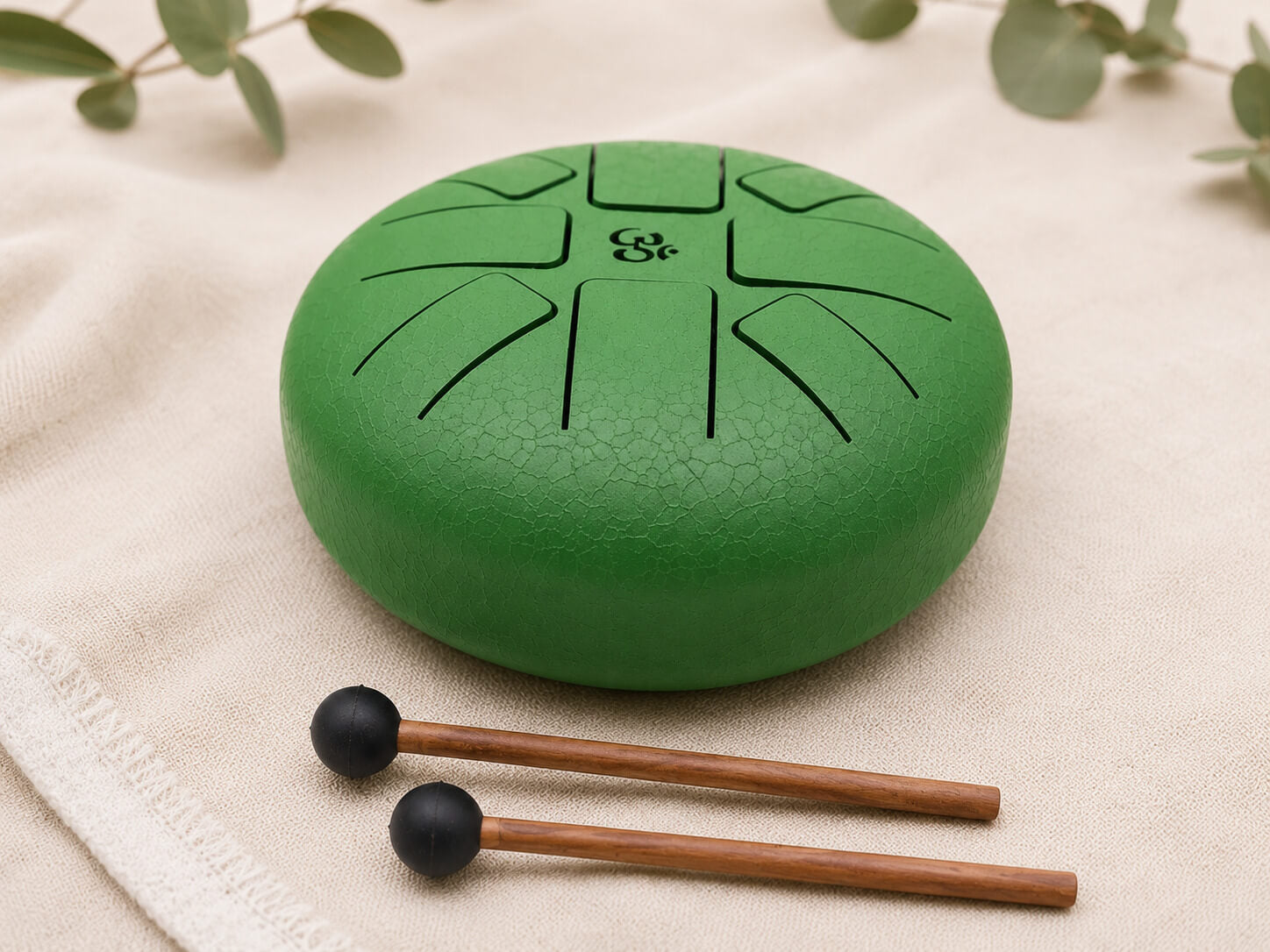 Tongue drum 16cm vert émeraude avec symbole Om gravé et deux mailloches en bois posées sur tissu écru.