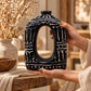Vase anneau ovale en terre cuite chocolat à motifs crème en relief, tenu entre deux mains devant un bouquet sec