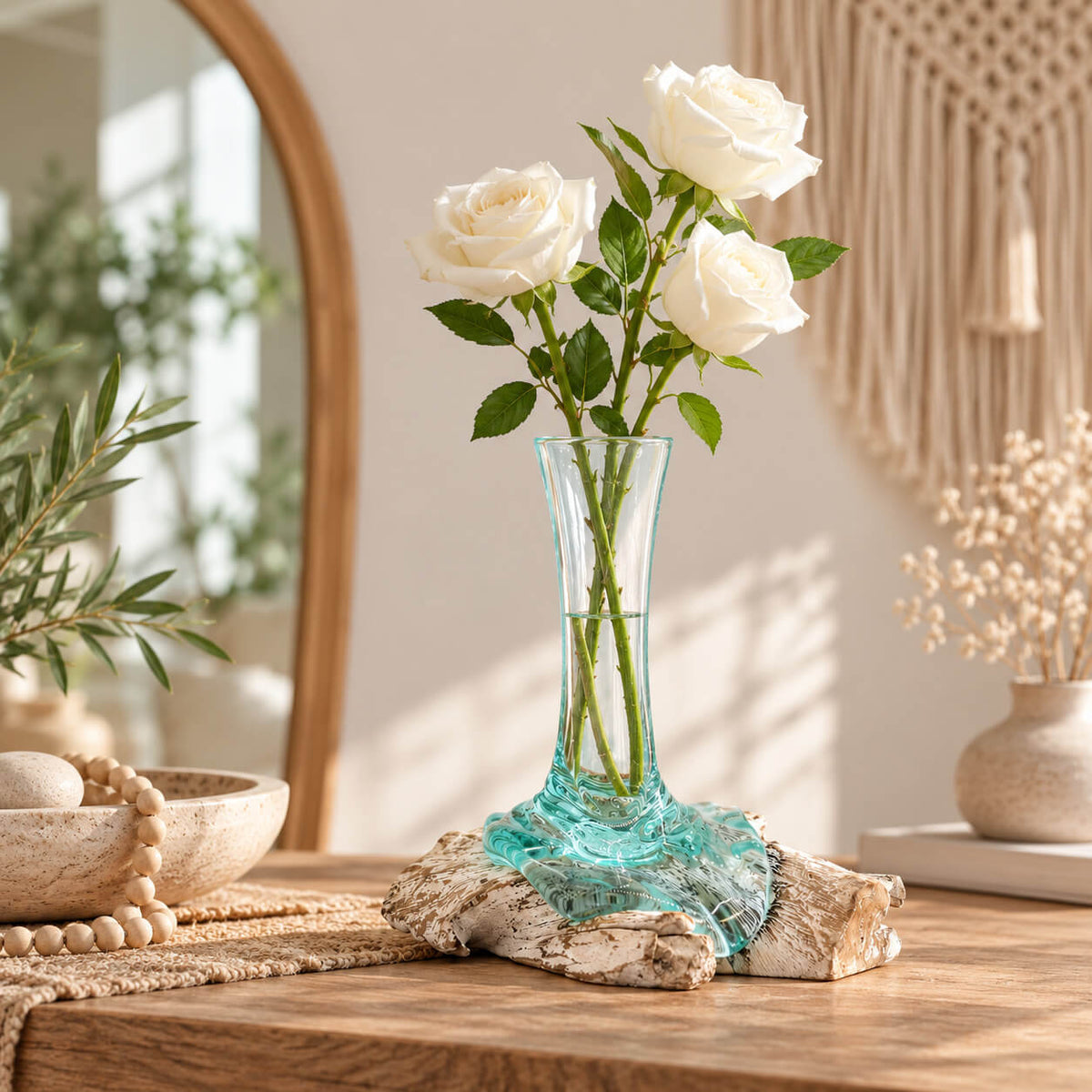 Petit vase en verre recyclé garni de trois roses blanches dans l'eau, sur sa racine blanchie posée sur une console en bois