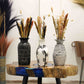 Trois vases décoratifs noir, blanc et gris contenant des bouquets de fleurs séchées, disposés sur une table en bois avec résine bleue.